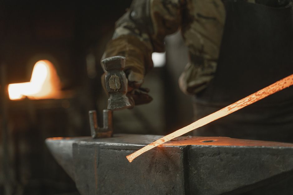 A blacksmith uses a hammer to shape hot iron on an anvil in a dimly lit workshop.