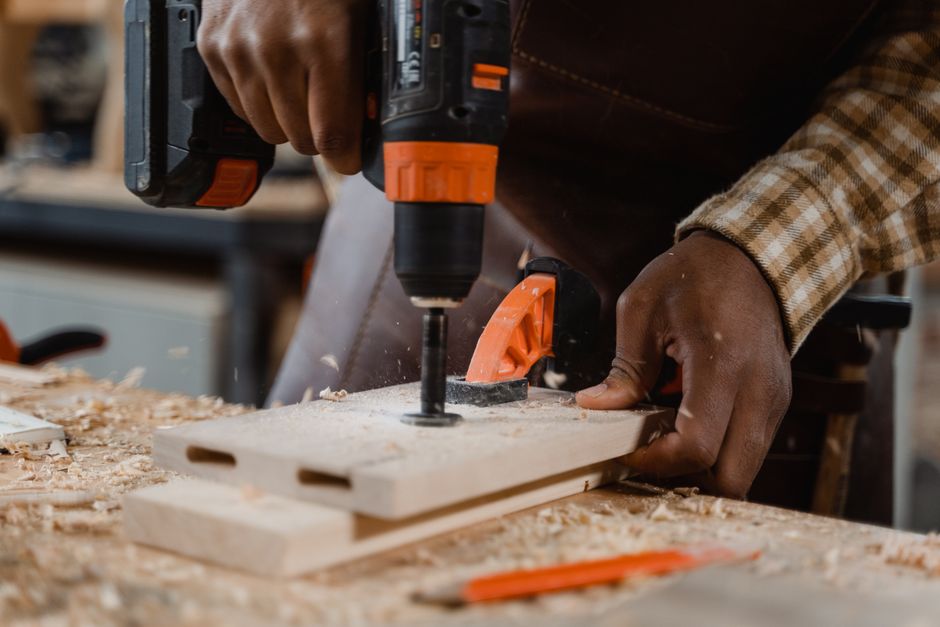 Carpenter uses drill on wood board, surrounded by sawdust in a workshop setting.